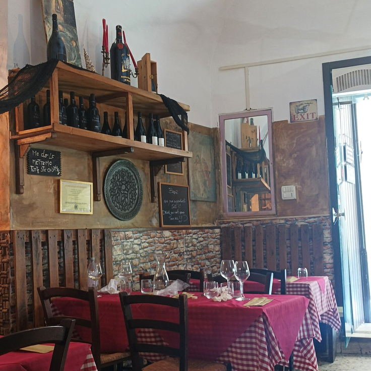 Angolo della sala con la scaffalatura del vino, una lavagnetta scritta a mano, diplomi incorniciati e un tavolo con la tovaglia a quadretti rossi.