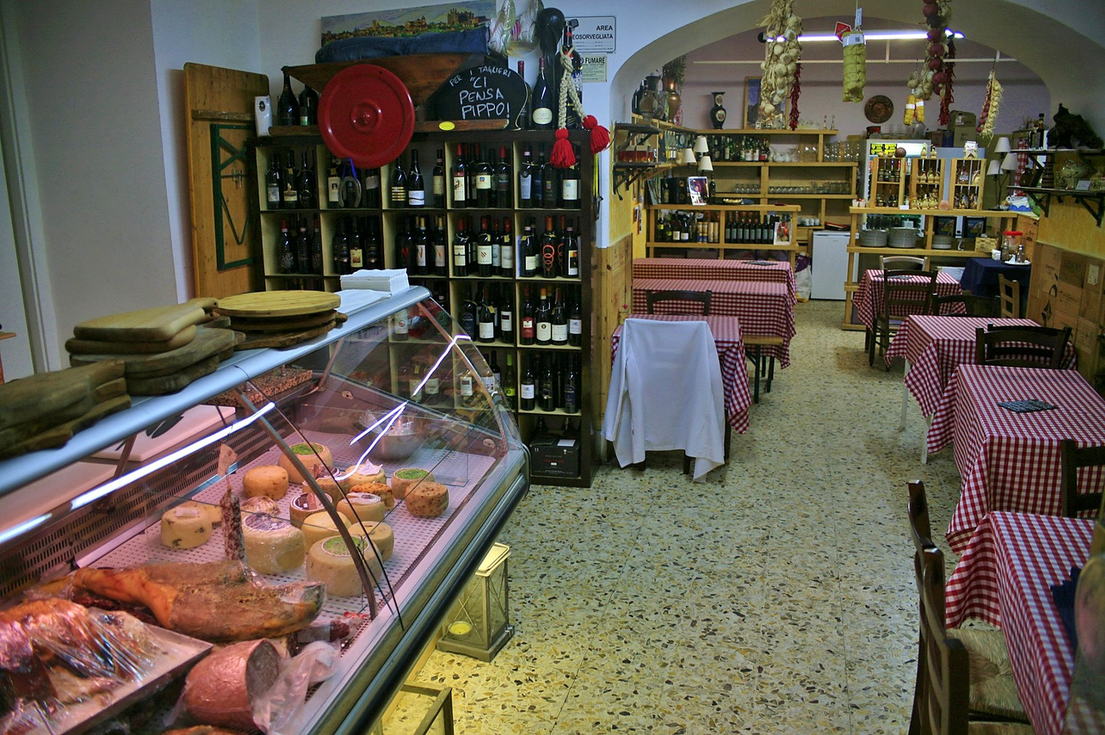 La sala vista dal banco: la vetrina dei salumi in primo piano, le bottiglie di vino sugli scaffali e i tavoli con le tovaglie a quadretti rossi.