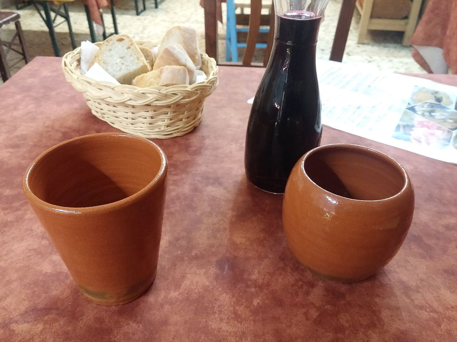 Due bicchieri di terracotta e una bottiglia scura di vino sfuso sul tavolo, con il cestino del pane.