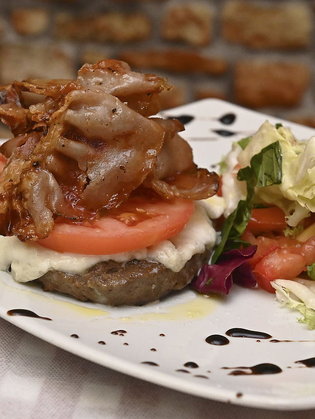 Hamburger di carne Chianina con cipolle stufate, servito con insalata e pomodoro, davanti al muro di mattoni.