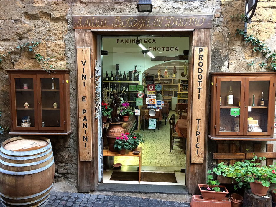 L'ingresso della bottega in Via Pedota: insegna dipinta a mano sopra la porta, due tavole in legno con le scritte VINI E PANINI e PRODOTTI TIPICI, una botte e i vasi di fiori.