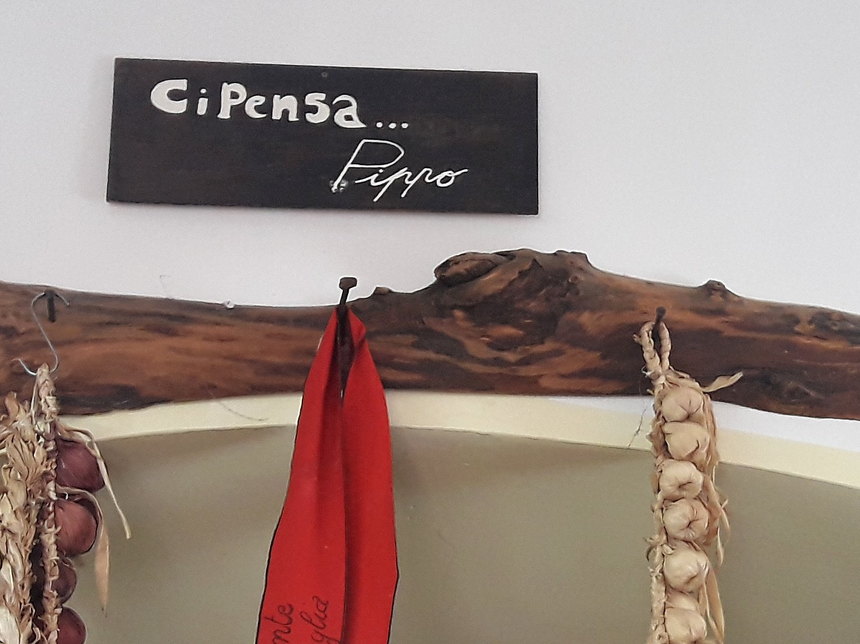 Lavagna appesa sopra una trave di legno con la scritta a mano «Ci Pensa... Pippo» e la firma; ai lati due trecce d'aglio e un panno rosso.