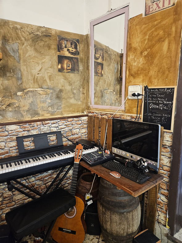 L'angolo del palco: pianoforte elettrico, chitarra appoggiata alla botte, mixer e la lavagna con l'annuncio dell'Open Mic.