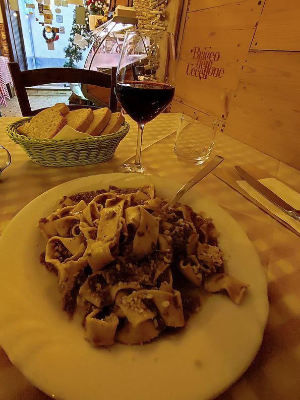 Piatto di pappardelle al ragù di cinghiale, con un bicchiere di vino rosso e il cestino del pane sul tavolo.