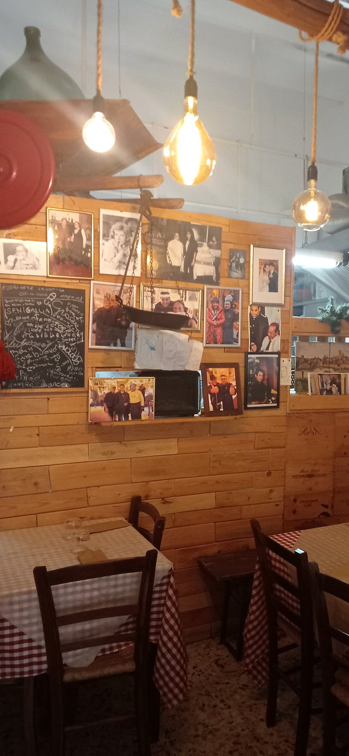 L'angolo della sala: la damigiana in alto, la vecchia bilancia appesa, la lavagna con le firme degli ospiti e la parete di fotografie sopra i tavoli a quadretti rossi.