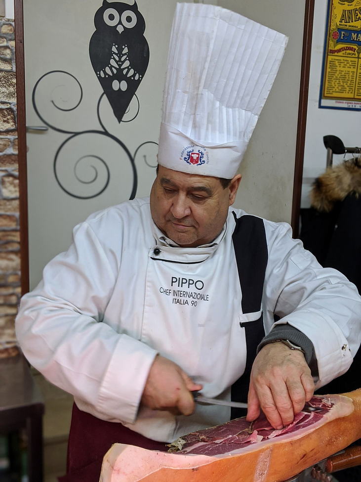 Pippo, in casacca bianca e cappello da cuoco, affetta un prosciutto crudo dietro il banco della bottega.