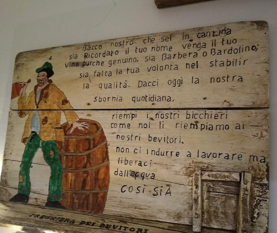 Tavola di legno dipinta e appesa in sala: un bevitore con il cappello verde accanto a una botte, e accanto il testo della «Preghiera dei Bevitori» scritto a mano.