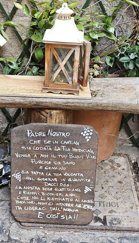 Tavola di legno appoggiata al muro di pietra accanto alla porta, con il testo del «Padre nostro del vino» scritto a mano in vernice bianca; sopra, una lanterna e l'edera.
