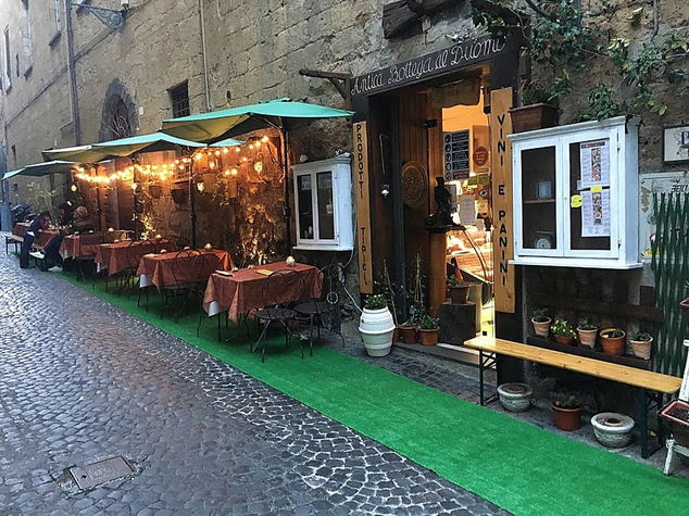 I tavoli all'aperto nel vicolo, la sera, con le luci a festone accese sopra la tenda della bottega.